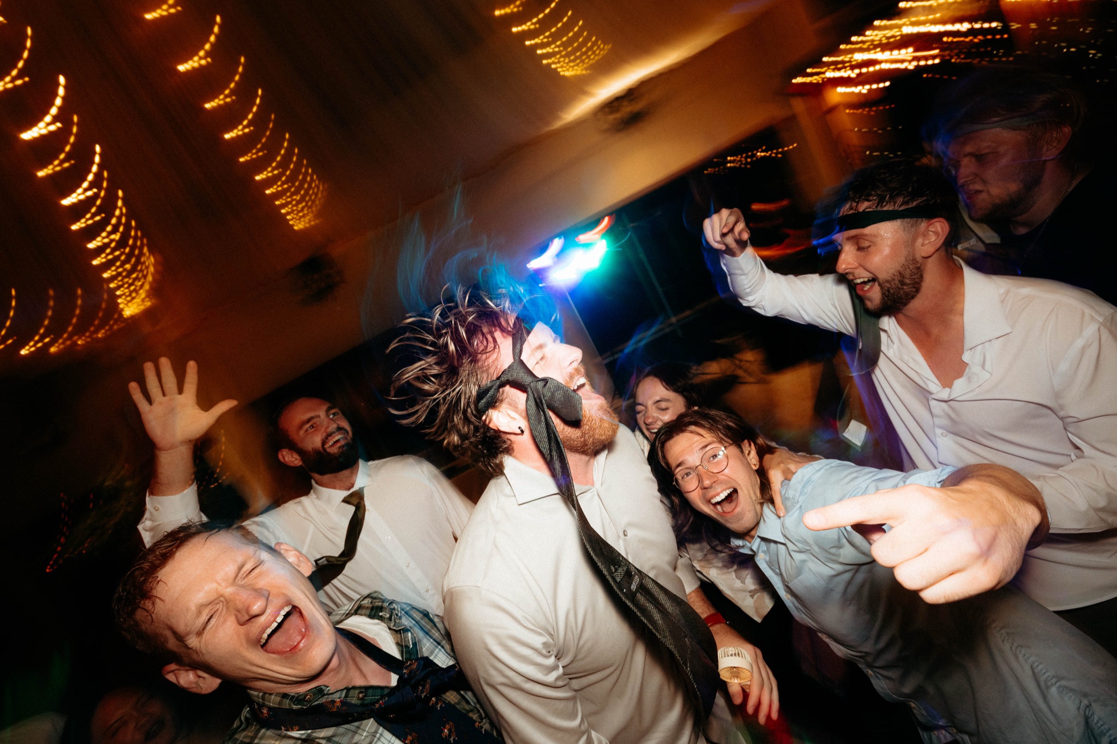 Guests laughing and dancing shoulder to shoulder under string lights, ties tied around their heads
